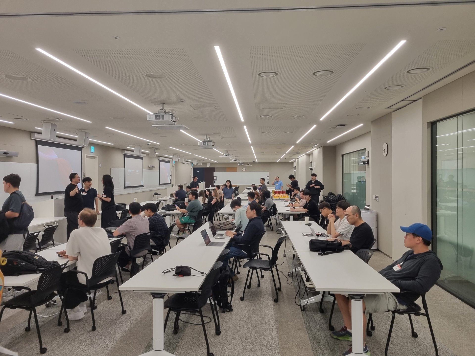 긴 테이블과 의자가 놓인 행사장에서 참가자들이 자리를 잡거나 서서 대화를 나누고 있다.